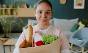 Young nurse holding paper bag full of healthy food while looking at camera and smiling widely