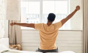 Arabic Man Sitting On Bed Back To Camera And Stretching Arms Awaking After Good Sleep In Modern Bedroom At Home, Looking Out Of Window. Comfort And Coziness. Good Morning Concept