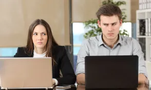 Front view of two angry businesspeople using computers disputing at workplace and looking sideways each other with anger