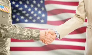 American soldier in uniform and civil man in suit shaking hands with national flag on background - United States