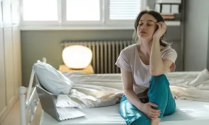 A young woman sitting on her bed in a sunny bedroom, appearing deep in thought and showing signs of tiredness. The scene suggests contemplation, relaxation, or a morning routine in a cozy setting.