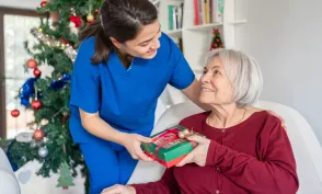 Home Caregiver Hugging And Giving Presents Senior Woman For Christmas At Nursing Home