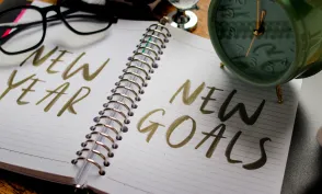 A notebook with the words NEW YEAR and NEW GOALS written on the pages, accompanied by a clock and a pair of glasses.