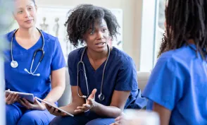 Nurses in blue scrubs sit together in a team meeting, discussing patient care while holding tablets.
