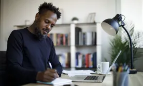 A student writes in a notebook at a desk with a laptop, phone, lamp, and bookshelves nearby.