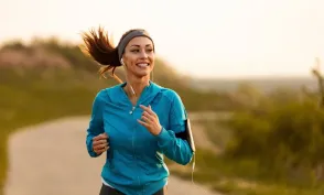 Woman smiling while jogging outdoors on a winding path, wearing earbuds and a blue jacket with a phone armband.