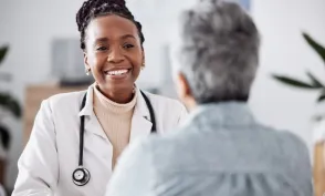 A smiling nurse practitioner wearing a white coat and stethoscope speaks with an older patient during an appointment.