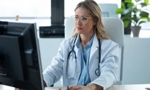 A nurse practitioner wearing a white lab coat and stethoscope sits at a desk using a computer in a bright medical office.