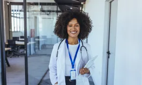 Smiling nurse practitioner in a white coat with a stethoscope holds a tablet in a modern medical office.
