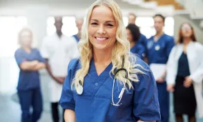 Smiling RN who is pursuing a BSN in blue scrubs with a stethoscope stands in the foreground while a multidisciplinary medical team gathers in the background of a healthcare facility.