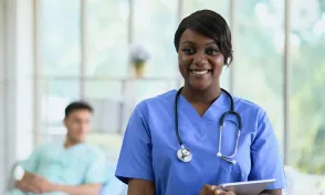 Smiling nurse in blue scrubs with a stethoscope and tablet standing in a bright hospital room, with a patient resting in a bed blurred in the background.