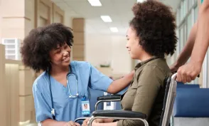 Registered nurse in blue scrubs speaks reassuringly with a patient seated in a wheelchair in a hospital hallway.
