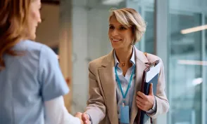 Professional shakes hands with a healthcare worker while holding a clipboard, suggesting a meeting or career opportunity in a healthcare setting.