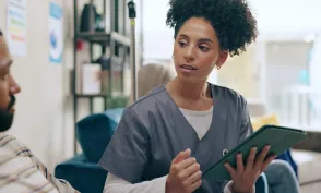Registered nurse who graduated from an MSN Direct Entry program in scrubs discusses information on a tablet with a seated man in a home setting.