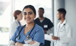 RN in blue scrubs with a stethoscope smiles confidently at the camera while colleagues collaborate in the background of a clinical setting.
