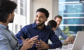 Four coworkers sit around a table in a bright office. A smiling man in a navy shirt talks with a colleague in the foreground while two others work near an open laptop in the background.