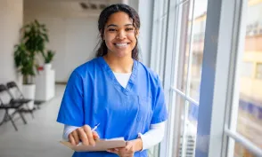 A smiling nurse in blue scrubs holds a clipboard and pen while standing beside large windows in a bright healthcare facility.