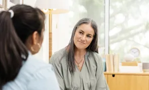 A holistic nurse listens attentively to a patient while holding a notepad and pen during a one-on-one conversation in a calm setting.