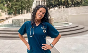 Herzing University online nursing student in blue scrubs and hands on hips smiling outside in courtyard