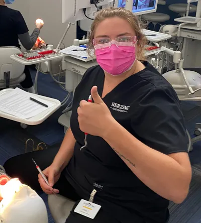 A Herzing dental hygiene student giving a thumbs up while performing a dental procedure. The student is wearing a pink mask and protective glasses, with a dental monitor showing images in the background. A patient is seated in the dental chair, receiving treatment.
