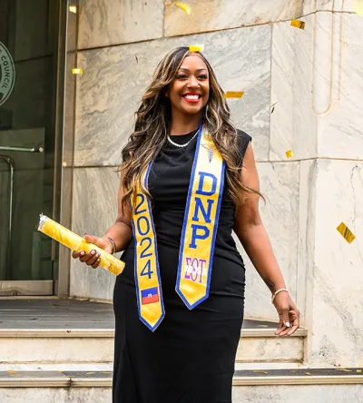 Graduating student celebrating with confetti, wearing a black dress and DNP 2024 sash, holding a diploma outside a building.