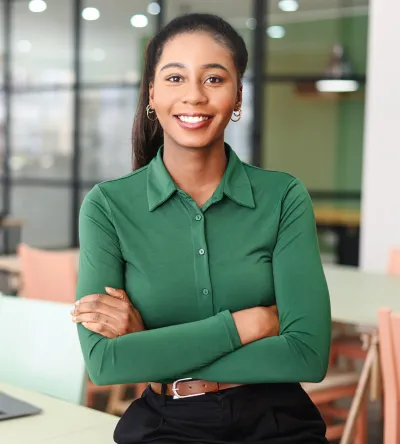 A professional woman in a green blouse sits at a modern office table, smiling with arms crossed, ready for career success.