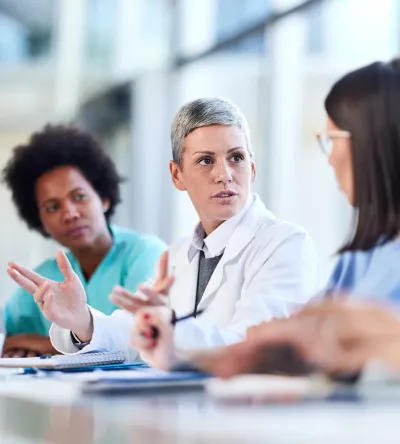 A medical leader in a white coat facilitates a collaborative team meeting with nurses and doctors in a hospital conference room.