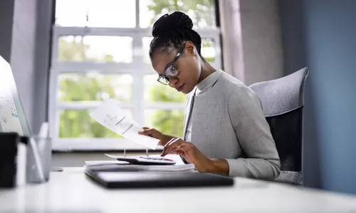 A Herzing University accounting student in professional attire reviews financial documents at a modern office desk, preparing reports and analyzing data.