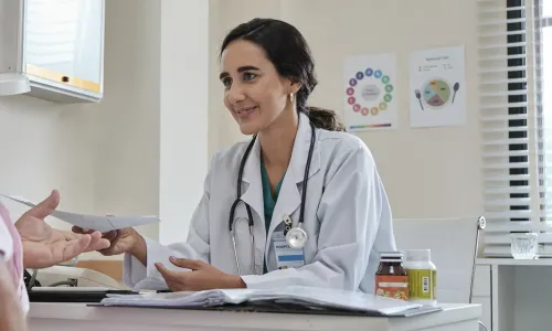 A female healthcare provider in a white coat and stethoscope smiles warmly while handing a paper to an older male patient during a consultation in a medical office A female public health nurse in a white coat and stethoscope smiles warmly while handing a paper to an older male patient during a consultation in a medical office