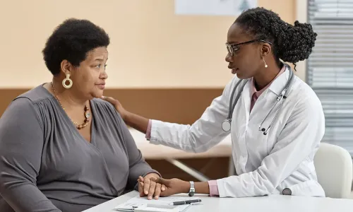 A nurse practitioner sits at a table with an adult patient, offering support by holding the patient's hand and placing a reassuring hand on her shoulder during a conversation in a medical office