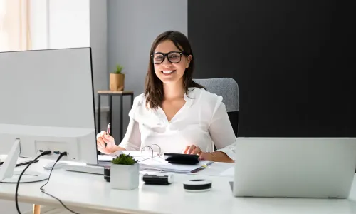 Smiling Texas woman in business casual attire working at a modern desk with dual monitors, calculator, and office supplies, in a bright and organized home or office setting