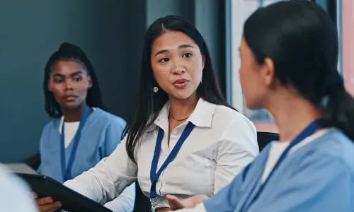 Three nurses, including two in scrubs and one with a doctoral degree wearing a lanyard, engage in a focused discussion in a professional or educational setting