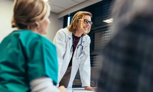 Smiling female healthcare educator in a white coat with a stethoscope leaning on a table, engaging with nursing students during a classroom discussion