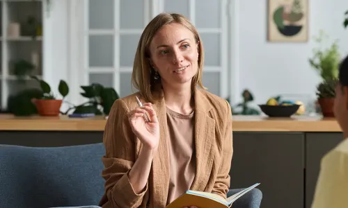 A social worker with a BSW attentively listens and takes notes while engaging in a one-on-one counseling session with a client in a comfortable, plant-filled office setting