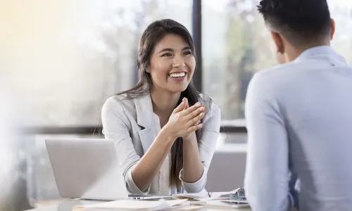Professional meeting between a smiling business professional and a client seated across a desk with a laptop and documents, engaged in conversation in a bright office setting.