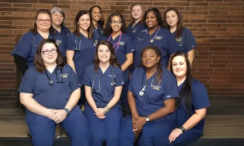 Group of LPN to RN students wearing blue scrubs smiling for picture in front of brick wall