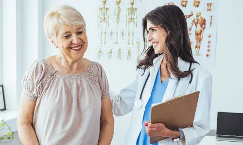 Smiling nurse wearing a white coat and stethoscope holding a clipboard, warmly interacting with an older female patient in a medical office Smiling women's health nurse practitioner wearing a white coat and stethoscope holding a clipboard, warmly interacting with an older female patient in a medical office