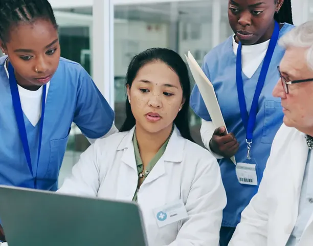 Medical team collaborating around a laptop, with two nursing students in blue scrubs and two healthcare professionals in white coats reviewing patient information