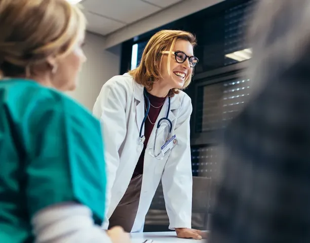 Smiling female healthcare educator in a white coat with a stethoscope leaning on a table, engaging with nursing students during a classroom discussion