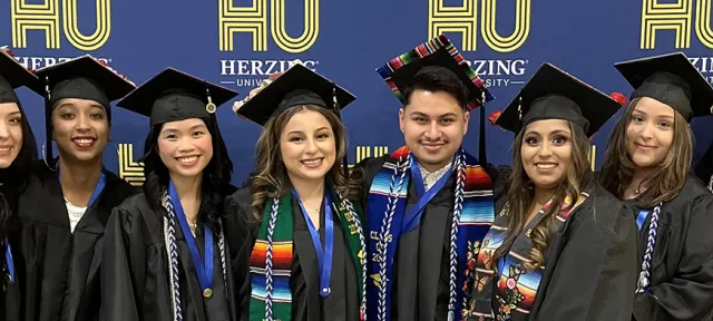 A group of Herzing University graduates in caps and gowns, smiling and posing together in front of a blue backdrop with the university’s logo.