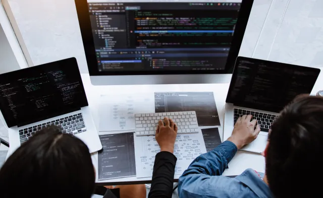 Two computer programmers seated at desk in front of multiple monitors showing coding software