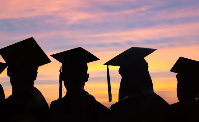 A group of seven nursing graduate students look of into the distance as the sun sets. They are wearing caps and gowns.