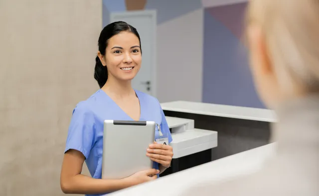 Medical assistant at front desk holding tablet smiling with patient