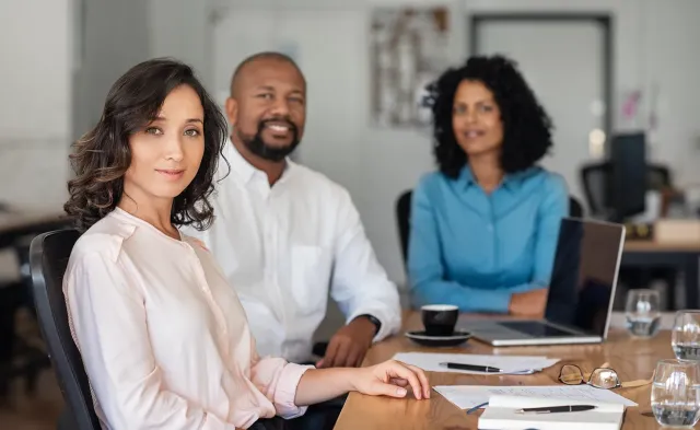 a group of three business professionals are setting in a board room. They are looking at the camera. 