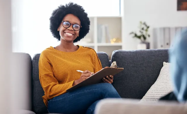 Smiling substance abuse counselor with natural hair and glasses sitting on a couch, holding a clipboard and pen during a therapy session.