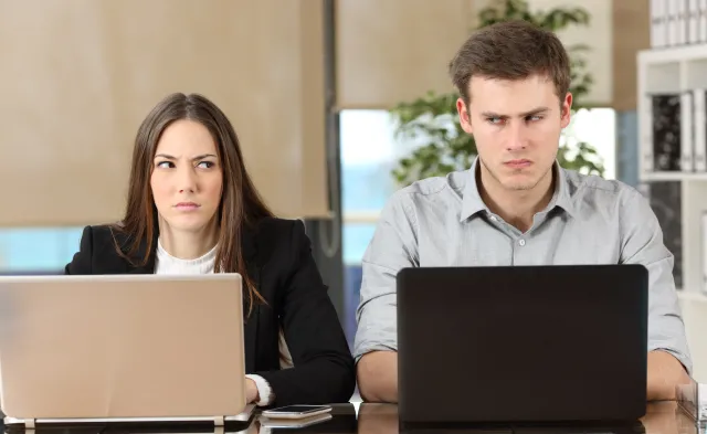 Front view of two angry businesspeople using computers disputing at workplace and looking sideways each other with anger