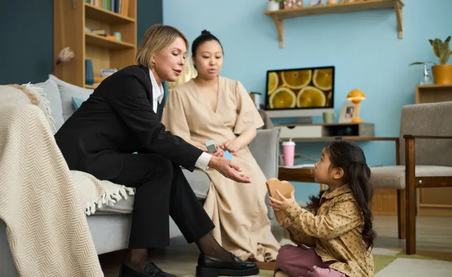 Caregiver sitting on couch, interacting with young child playing with toys, young mother observing in background