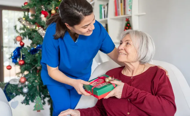 Home Caregiver Hugging And Giving Presents Senior Woman For Christmas At Nursing Home