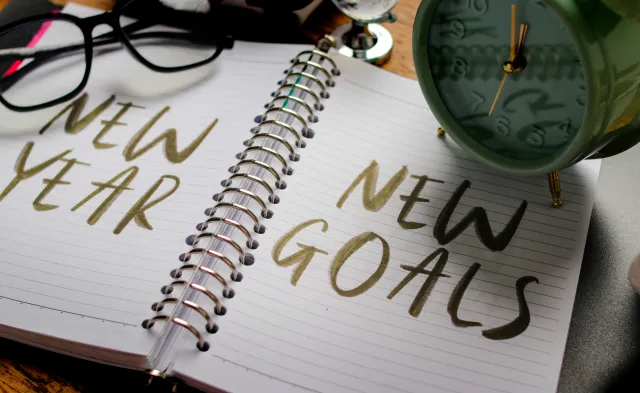 A notebook with the words NEW YEAR and NEW GOALS written on the pages, accompanied by a clock and a pair of glasses.