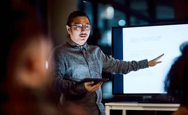 IT project manager presenting information on a large screen while holding a tablet and speaking to colleagues in a modern office.